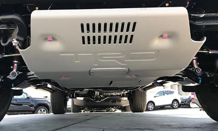 Underside view of a vehicle showing a light grey TRD skid plate with visible bolts, surrounded by suspension components.