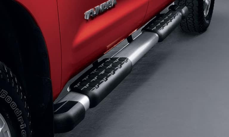 An aluminum running board with black textured step pads installed on the side of a red Toyota Tundra.