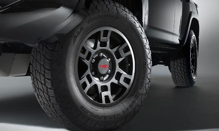 Close-up of a dark SUV's front wheel, showcasing the rugged all-terrain tire and silver multi-spoke rim with a red TRD cap.