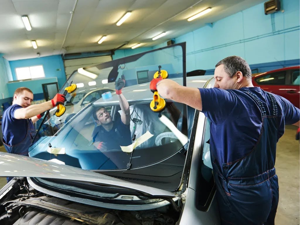 Two auto technicians installing a new windshield in a car using suction cups at a garage.