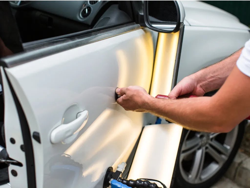 A person's hands performing paintless dent repair on a white car door, illuminated by two yellow light ramps.
